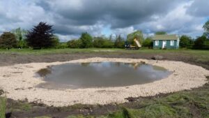 Village pond under contruction with digger in distance next to green and white Reading Room