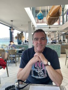 Man in blue t shirt sitting in restaurant with sea in back ground.