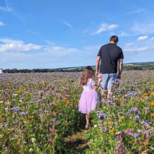 man walking along path in wild flower meadow, followed by little girl in a pink dress