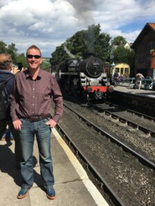 Man smiling next to a red and black steam train, chugging into station.