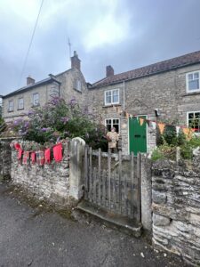 Cottage with bunting decorating stone walls near gate and scarecrow in garden. 