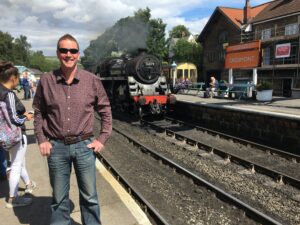 Man in jeans and sunglasses smiling at camera with steam train behind him