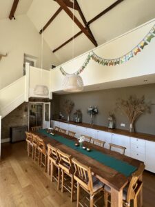 Dining room with high ceiling and beams. Wooden table with 14 chairs. Birthday bunting hung above. 