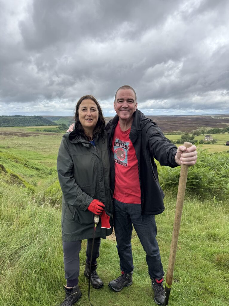 A man in a red t-shirt and black jacket, holding a tool, stands next to a woman in a green jacket and red gloves, both smiling at the camera in a grassy, hilly landscape under a cloudy sky.