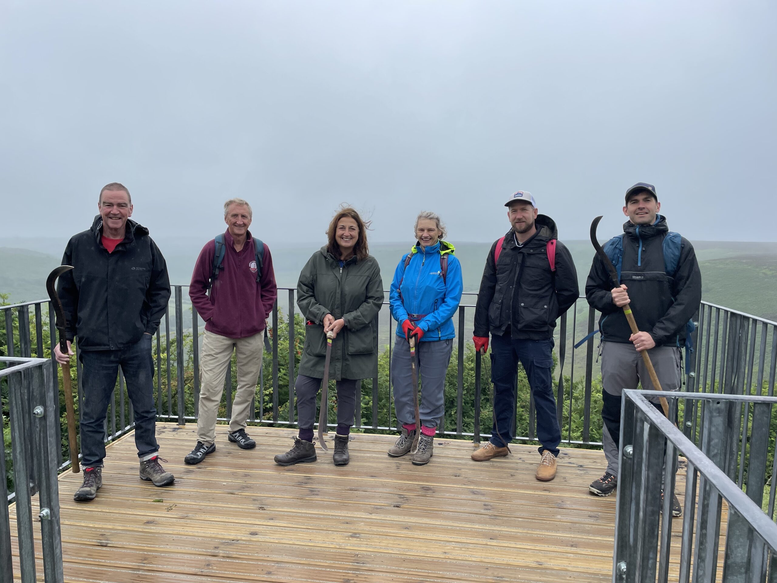A group of six smiling people stand on a wooden viewing platform, holding tools resembling slashing hooks. They are dressed in outdoor gear, including jackets, hiking boots, and hats, under a cloudy sky with a treeline visible in the background.