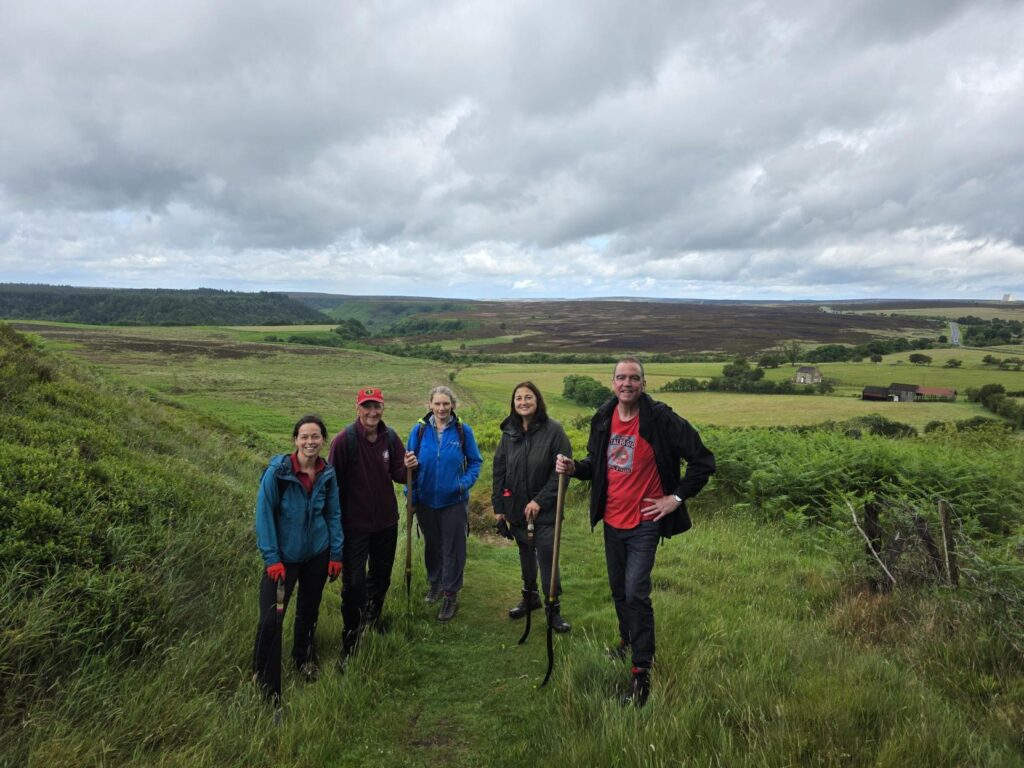 Five people, dressed in outdoor clothing and holding tools, are standing on a grassy path with a vast, rolling landscape of fields, hills, and a cloudy sky behind them.