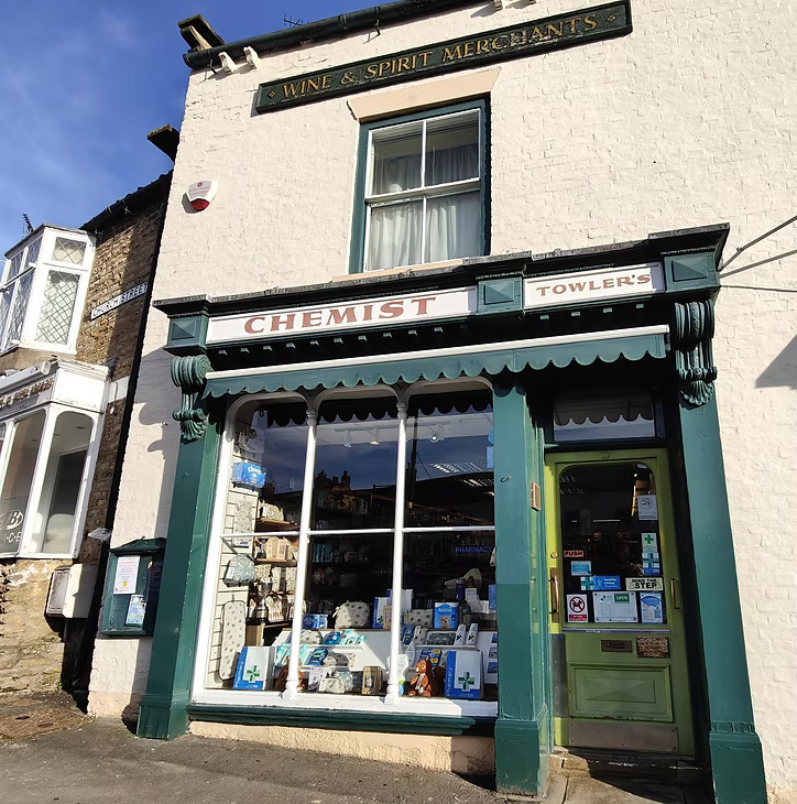 A slightly angled exterior view of a traditional shop building made of light-colored brick with a prominent dark green storefront. The shop's facade features a large display window filled with various products, and an ornate sign above reads "CHEMIST" on the left and "TOWLER'S" on the right. Above the first-floor window, a sign indicates "WINE & SPIRIT MERCHANTS." To the left, part of an adjacent building with a bay window and a street sign for "SMIDDY STREET" is visible. A light green door on the right of the storefront has a push bar and various notices.