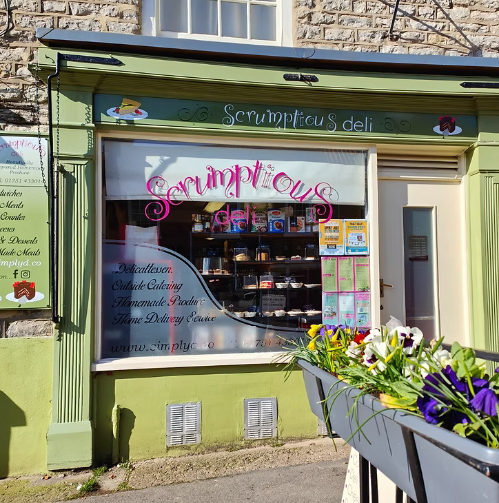 A full exterior shot of "Scrumptious Deli," a quaint shop with a green and light green facade. The shop window, adorned with pink cursive lettering of its name, displays various baked goods and products. Signs in the window advertise "Delicatessen," "Outside Catering," "Homemade Produce," and "Home Delivery Service." A menu is visible to the left of the window, and a flower box with colorful pansies and daffodils sits in the foreground.