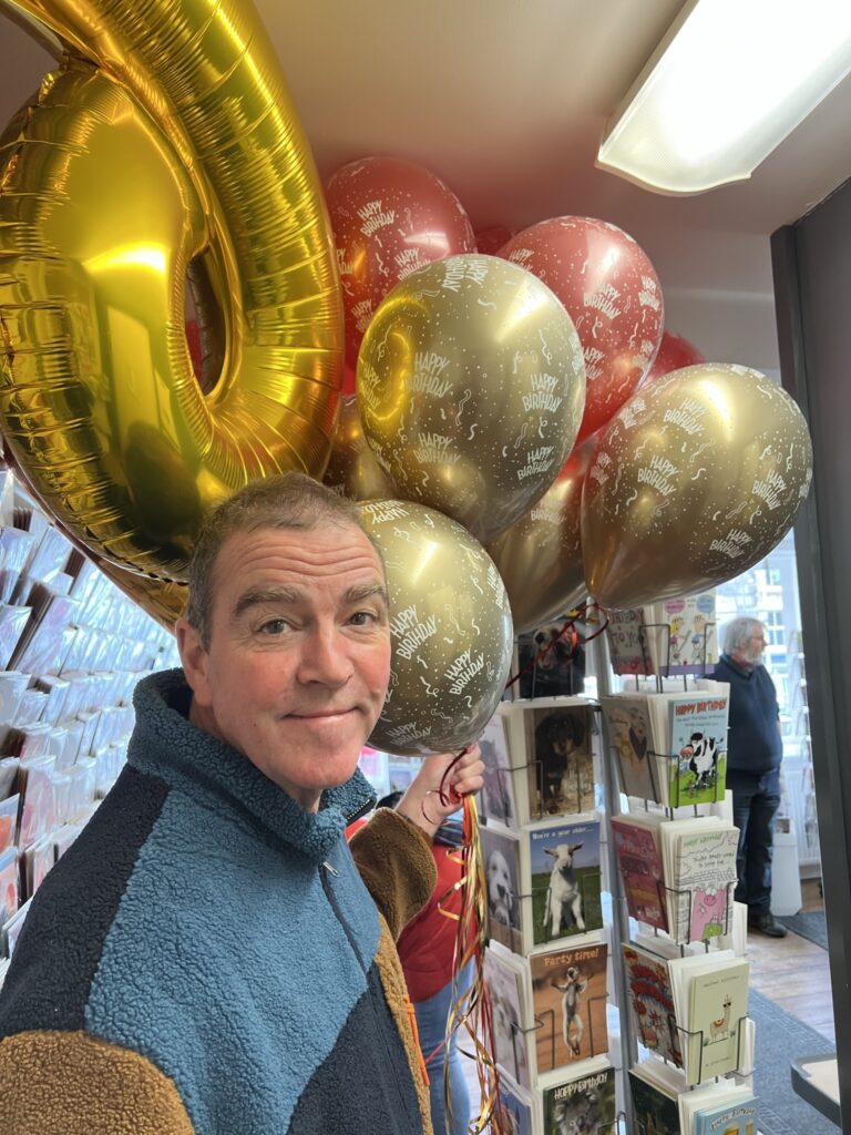 A candid, medium close-up shot of a smiling man with short, light brown hair, looking towards the camera. He is wearing a blue and brown fleece jacket. Behind and to the right of him is a large cluster of balloons, including a prominent gold "6" shaped balloon and several gold and red balloons that say "Happy Birthday." To his right, there are racks of greeting cards, and in the background, another person is partially visible. The setting appears to be inside a shop.