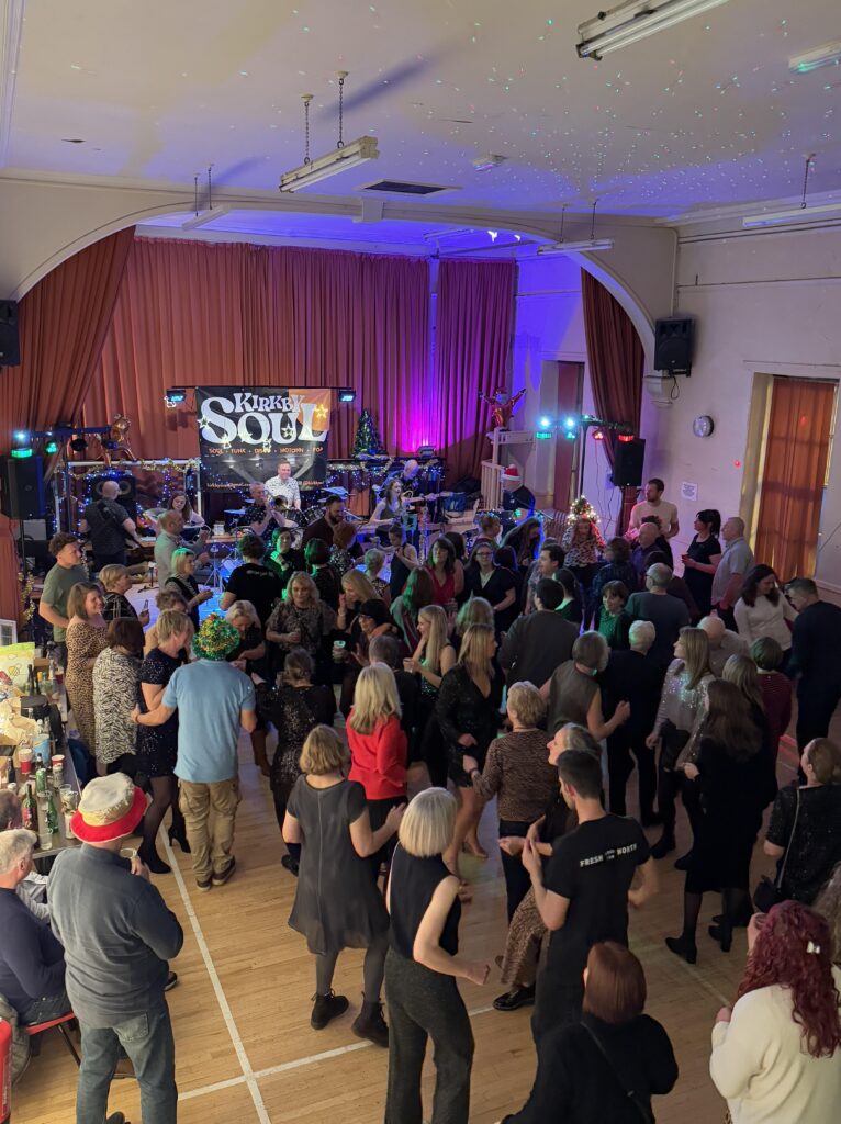 A vibrant, elevated view of a large gathering of people dancing and socializing in a spacious hall. A band is performing on a stage at the far end of the room, with a prominent "Kirkby Soul" sign visible. The stage is lit with colorful lights, and there are various band members and equipment visible. The floor is wooden, and there's a long table with drinks on the left side. The attendees are dressed in various outfits, and the overall atmosphere appears festive.
