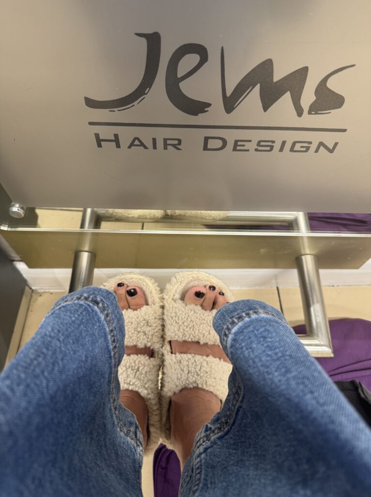A first-person, eye-level shot looking down at a person's feet, clad in light-colored, fluffy open-toed slippers, with dark painted toenails. The person is wearing blue jeans. In the background, above their feet, is a sign that reads "Jems" in a cursive font, with "HAIR DESIGN" in a sans-serif font below it. The setting appears to be a hair salon, with a metallic footrest visible.