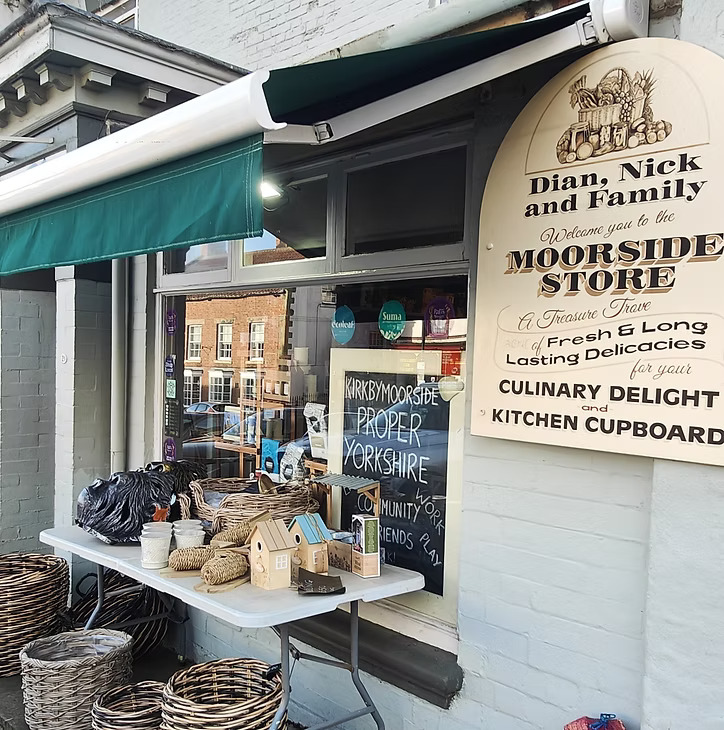 An exterior shot of a shop called "Moorside Stores," with a green awning extended over a display table outside. The table holds various woven baskets, small decorative birdhouses, and other household items. To the right of the window, a large, cream-colored wooden sign welcomes customers to "Moorside Stores," describing it as "A Treasure Trove" and a source of "Fresh & Long Lasting Delicacies" for "CULINARY DELIGHT and KITCHEN CUPBOARD." Inside the window, a sign reads "KIRKBYMOORSIDE PROPER YORKSHIRE."