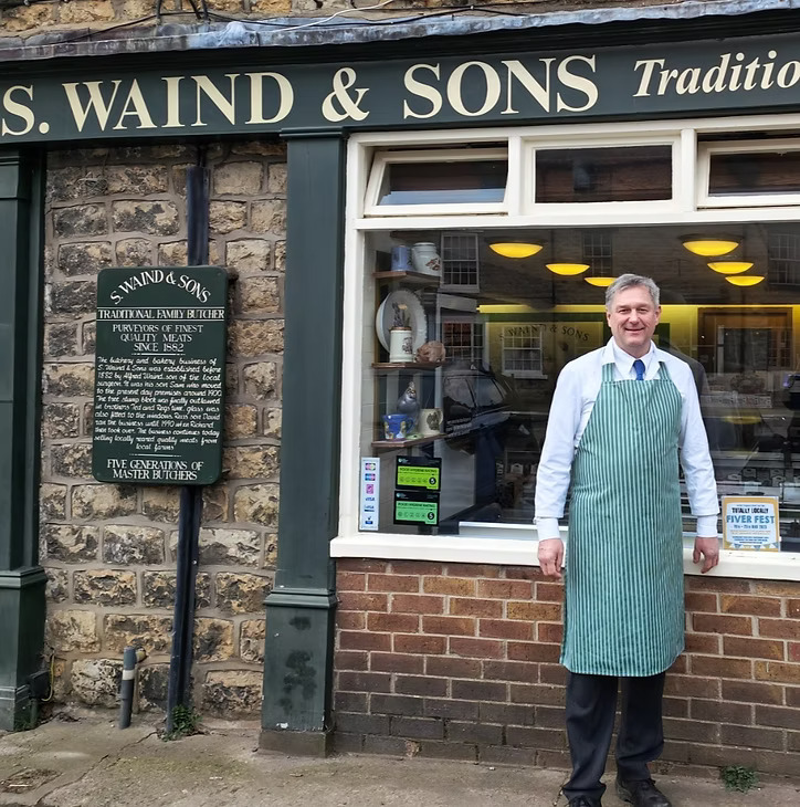 A smiling man, wearing a light blue striped butcher's apron over a white shirt and tie, stands in front of a traditional brick butcher shop with a dark green storefront. The shop's name, "S. WAIND & SONS Traditional Family Butcher," is prominently displayed above the window. To the left of the man, a dark green plaque on the stone wall details the history of the S. Waind & Sons establishment, mentioning "FIVE GENERATIONS OF MASTER BUTCHERS." The shop window displays various items, and a smaller sign inside reads "S. Waind & Sons."