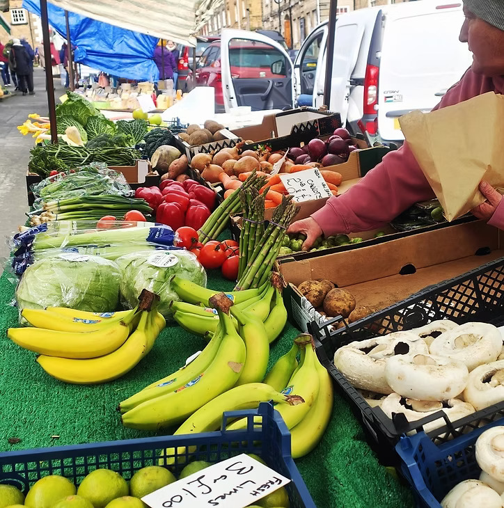 A bustling market stall laden with a vibrant array of fresh produce. In the foreground, large bunches of yellow and green bananas are prominently displayed on a green surface, next to crates of white button mushrooms and green limes. Behind them, a colorful selection of vegetables includes bright red tomatoes, green asparagus, celery, onions, potatoes, and various leafy greens. A person's hands are visible on the right, reaching for produce and holding a brown paper bag, suggesting a transaction is taking place. In the background, other market stalls and vehicles are partially visible, under what appears to be a clear sky.