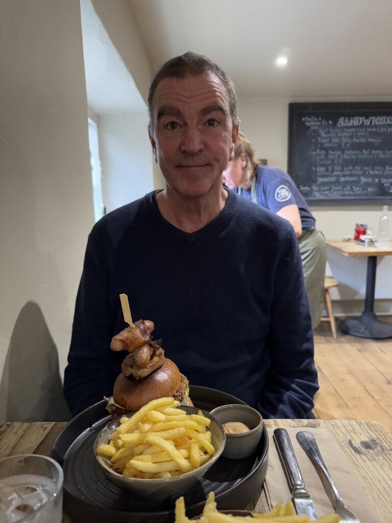 A man with short brown hair, a blue sweater, and a relaxed smile is seated at a wooden table in a cafe, looking directly at the camera. In front of him is a tall burger with a skewer holding bacon and other toppings, a bowl of french fries, and a small dish of sauce. In the background, another person is partially visible, leaning over a table. A chalkboard menu is on the wall behind them.
