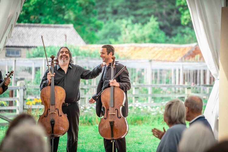 Two cellists, smiling, stand on a grassy lawn with their instruments. One man has his hand on the other's shoulder. An audience, including one person clapping, is visible in the foreground. In the background, there is a fence, trees, and buildings.