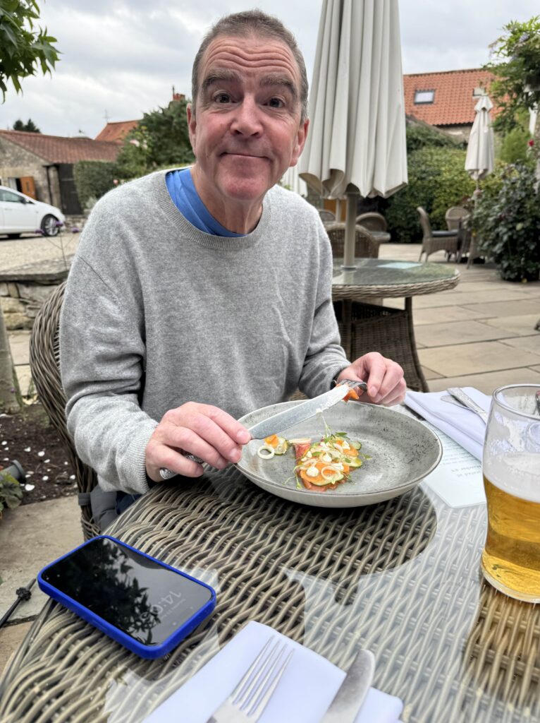 A person with short brown hair, wearing a light gray sweater over a blue shirt, is seated at a wicker table outdoors. They are smiling and looking at the camera while holding a fork and knife over a plate of food. A glass of beer and a phone with a blue case are on the table. The background shows a building, and there are white umbrellas in the distance.