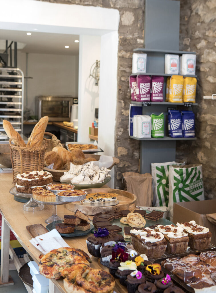 A close-up shot of a rustic wooden table piled high with a superb selection of baked goods. In the foreground, there are cupcakes decorated with icing and edible flowers, as well as slices of different cakes and brownies. In the background, there is a large wooden bread trough filled with baguettes and croissants, and on a shelf against a stone wall, there are bags of organic flour in various colors.