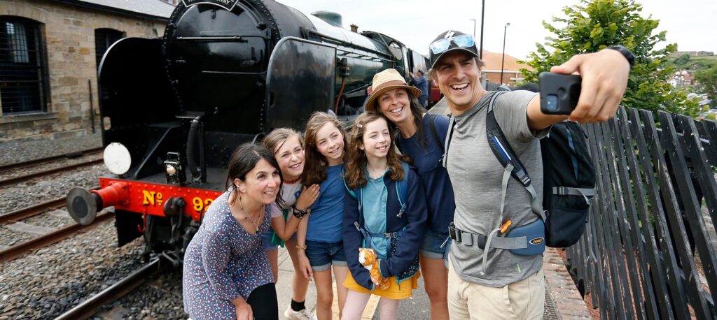 A man takes a selfie with a group of people, including two women and three children, standing on a train platform next to a black steam locomotive.