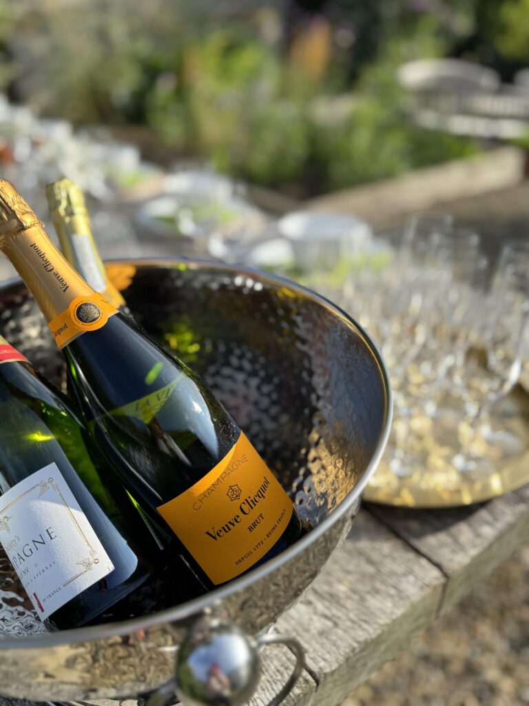 This image captures a celebratory outdoor setting, likely a garden party or reception, focusing on beverages. In the foreground, there is a large, textured silver ice bucket holding several bottles of sparkling wine or champagne. The most prominent bottle is a Veuve Clicquot Brut Champagne, easily identifiable by its signature bright yellow/orange label. A portion of another bottle, a white label with red text, is also visible. The ice bucket is resting on a weathered, light wooden surface, possibly an outdoor table or bench. In the background, slightly out of focus, there are several champagne flutes arranged on a gold, reflective tray. The area beyond that is a bright, sunny garden, with soft-focus greenery, flowers, and a natural, relaxed atmosphere. The lighting suggests it is a clear day.