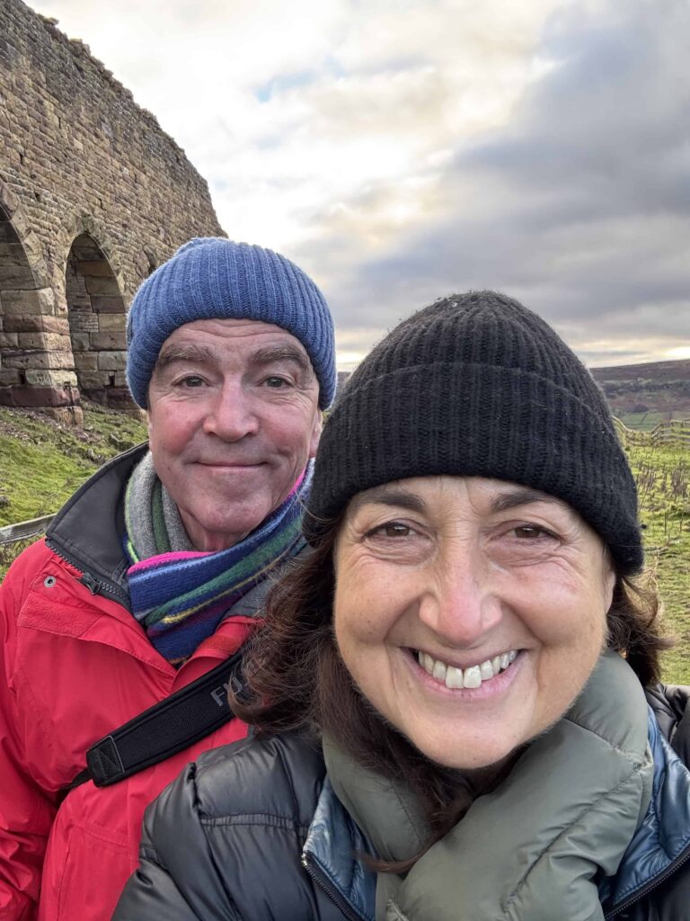  A smiling man and woman wearing winter hats and jackets take a selfie in front of arched stone ruins and a rolling green landscape under a cloudy sky.
