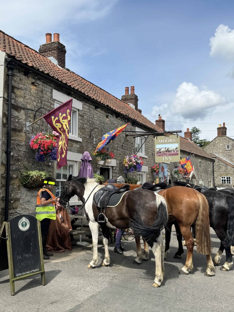 Several saddled horses stand on a road in front of "The Moors Inn," a stone building decorated with colorful flags and hanging flower baskets.